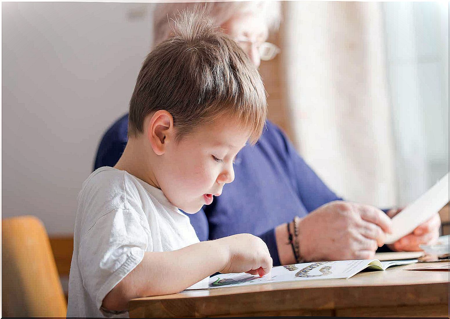 A small child reads at a table next to his grandmother